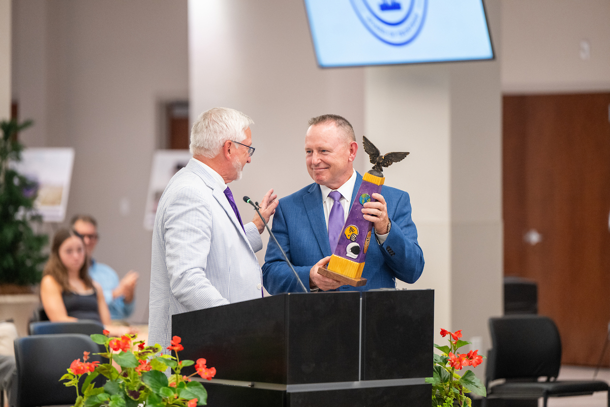 Wilmore holding an award topped by an eagle and smiling at Oldham.