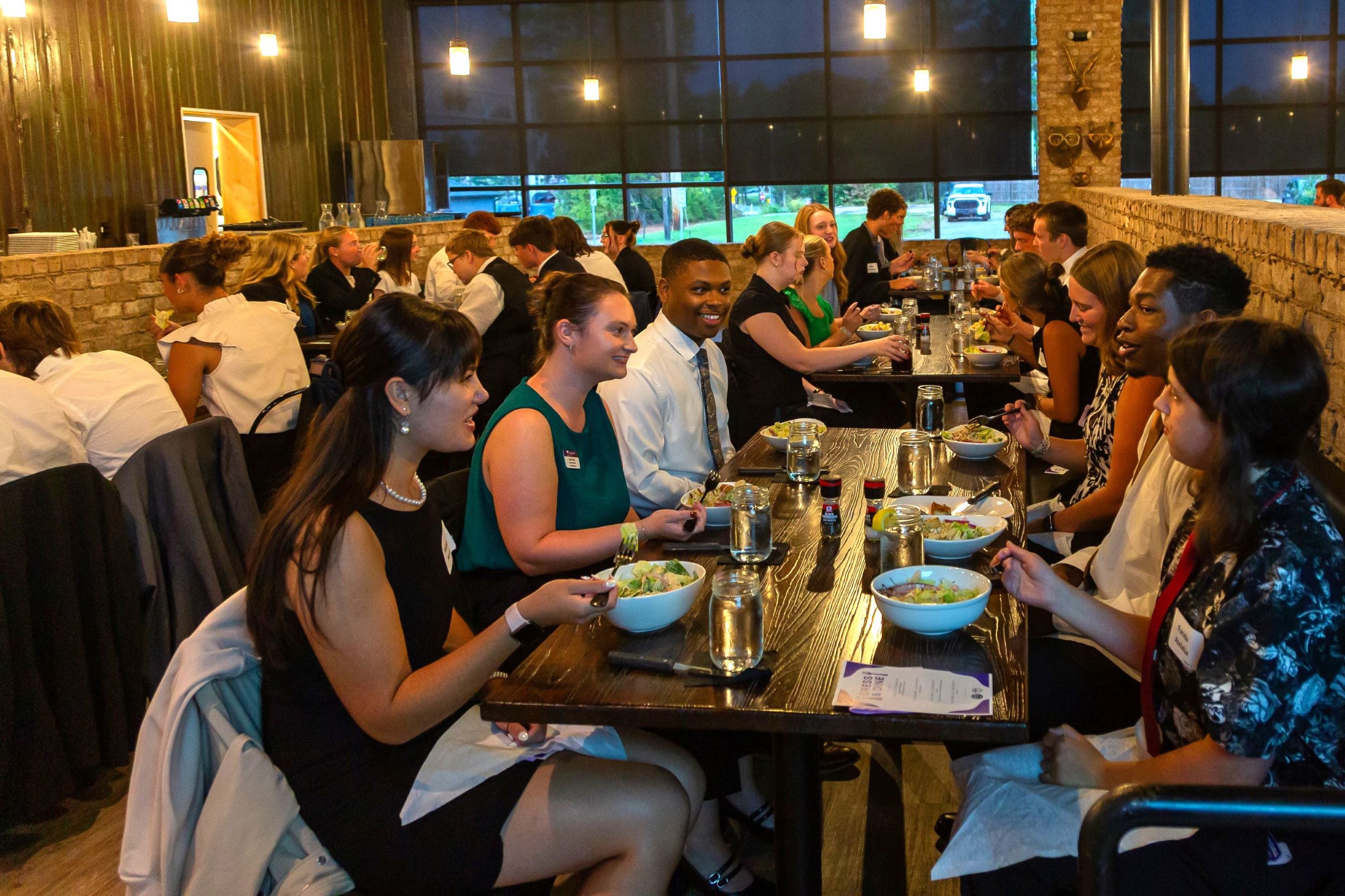 Students and professionals sit at long tables sharing a meal and conversation during a networking dinner inside a restaurant.