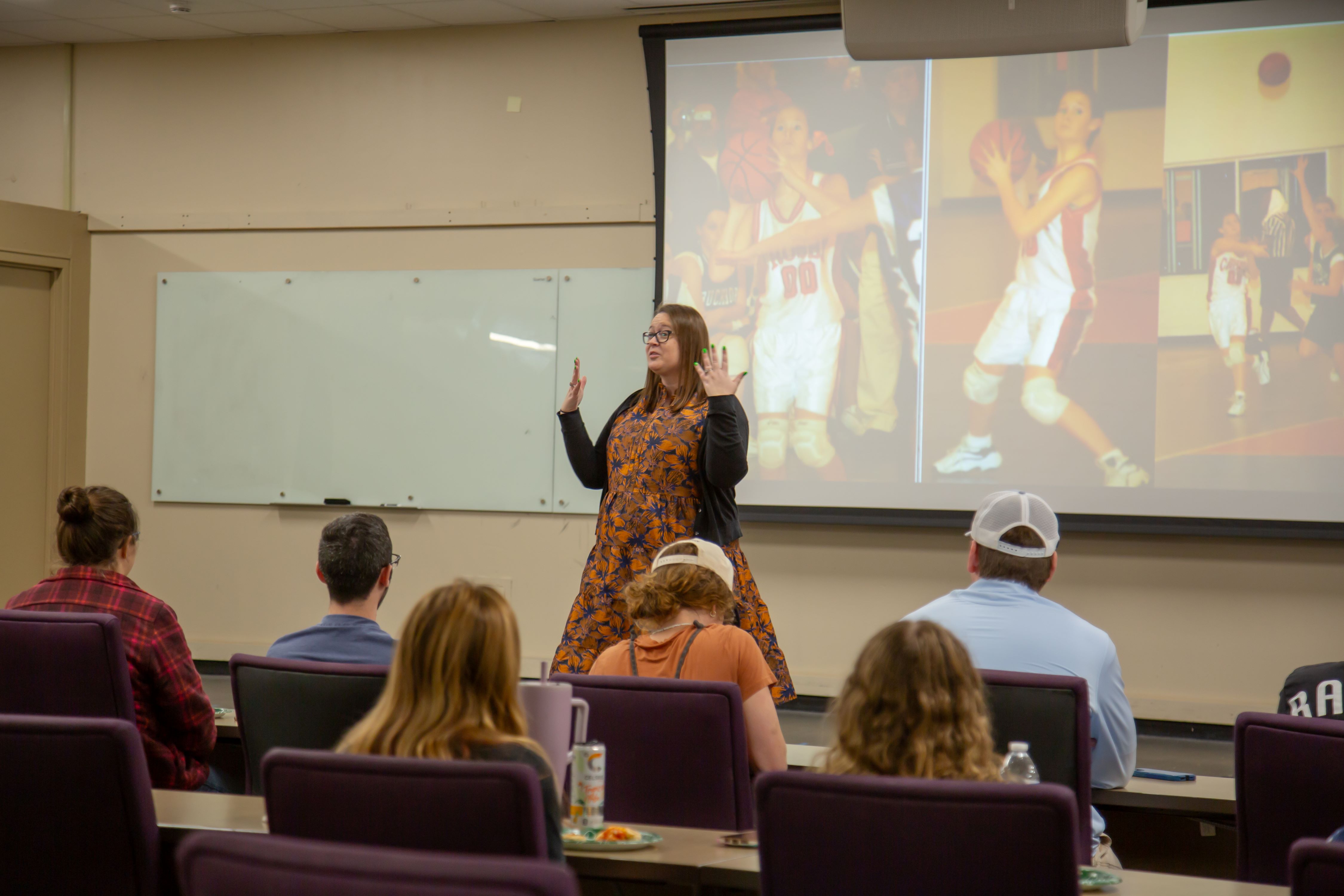 Dr. Chelsea Dowell gestures while presenting to students seated in a classroom, with a projected slide behind her showing basketball photos.