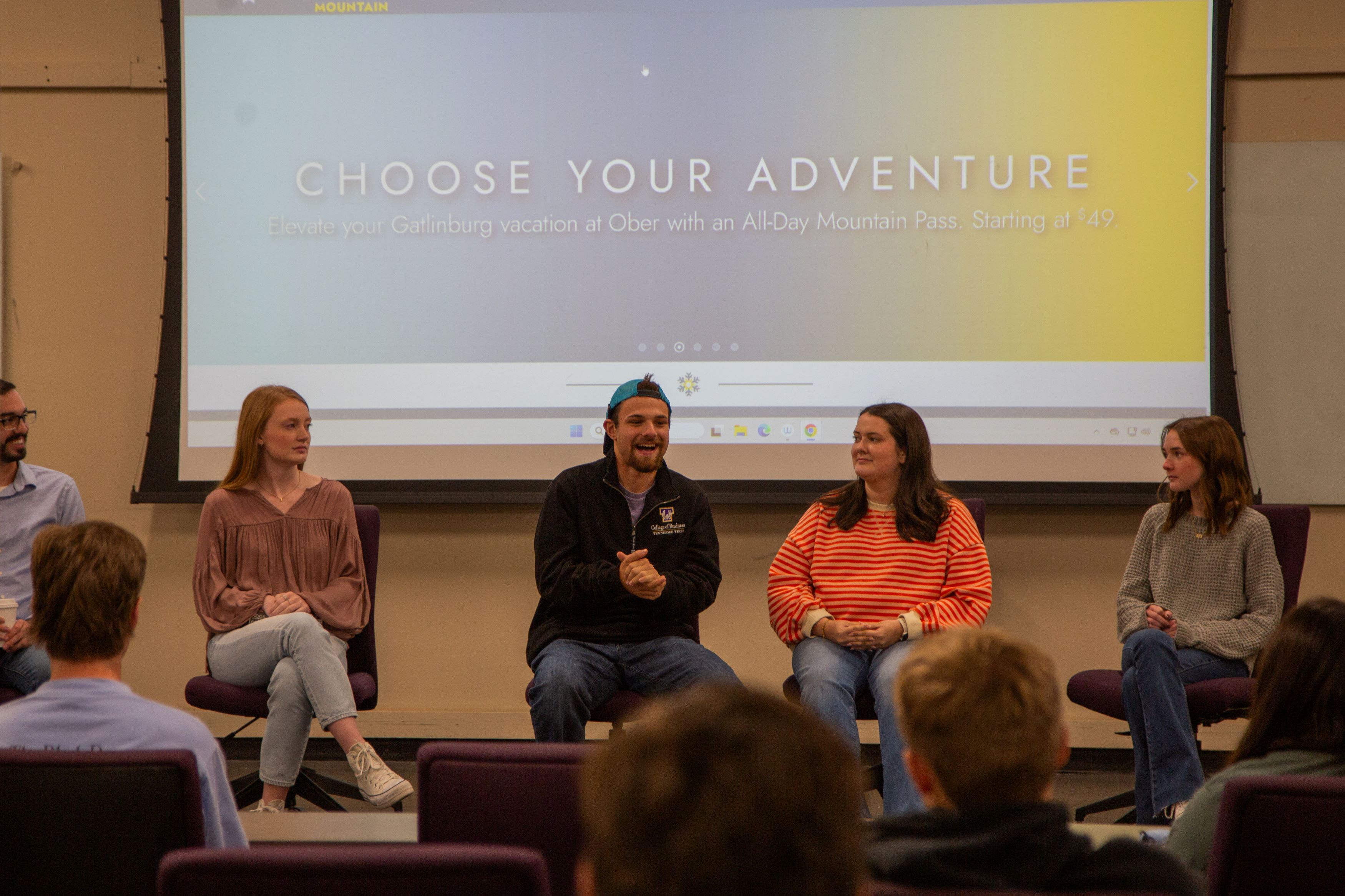 Five panelists sit at the front of a classroom speaking to an audience, with a projected screen behind them reading “Choose Your Adventure.”