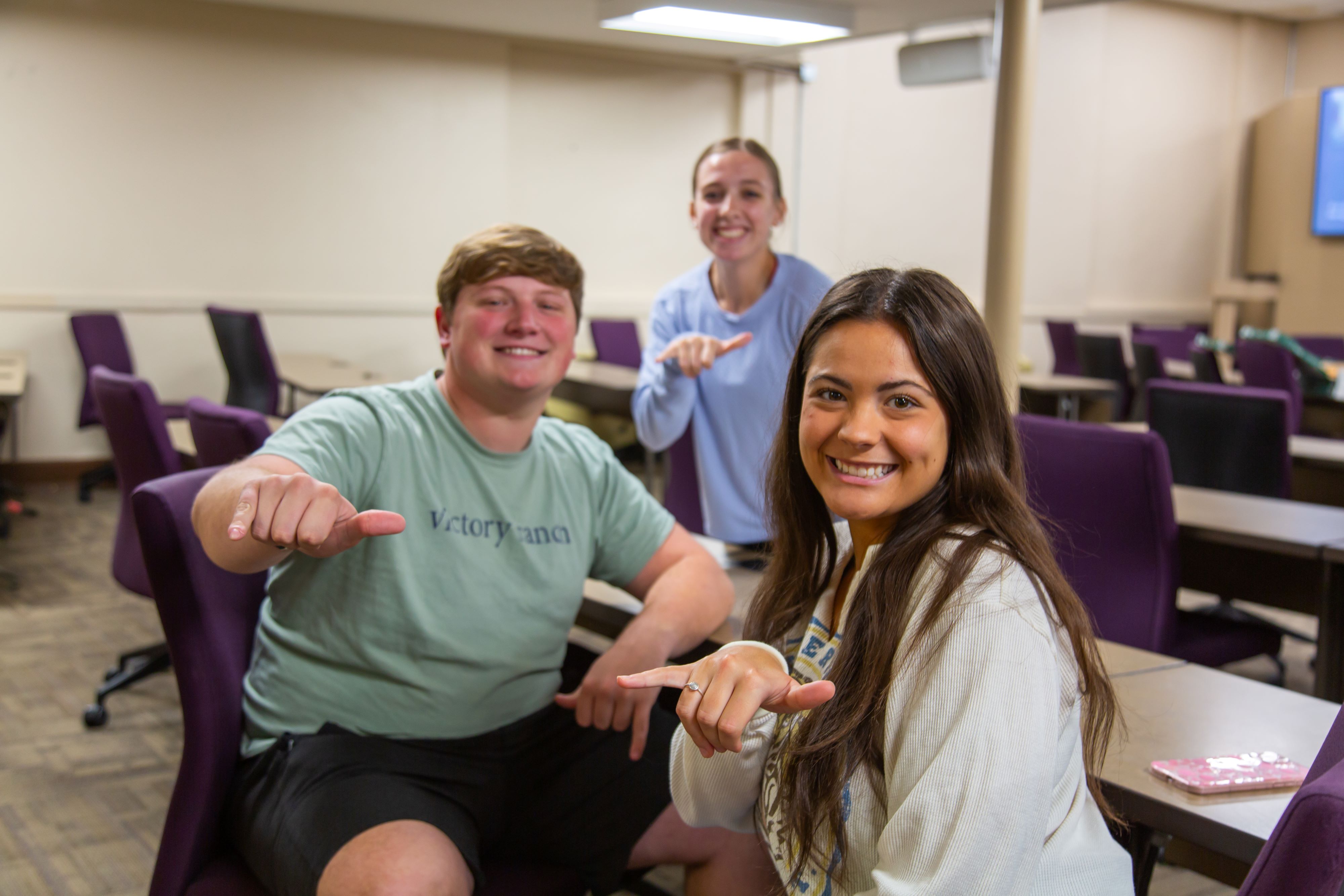 Three students smile and gesture toward the camera while seated in a classroom with purple chairs and tables.