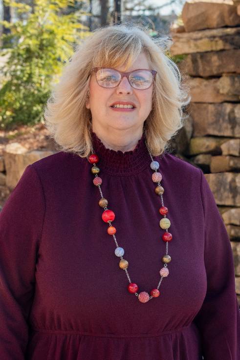 Female blond Caucasian wearing glasses and a dress with a long neckless. 