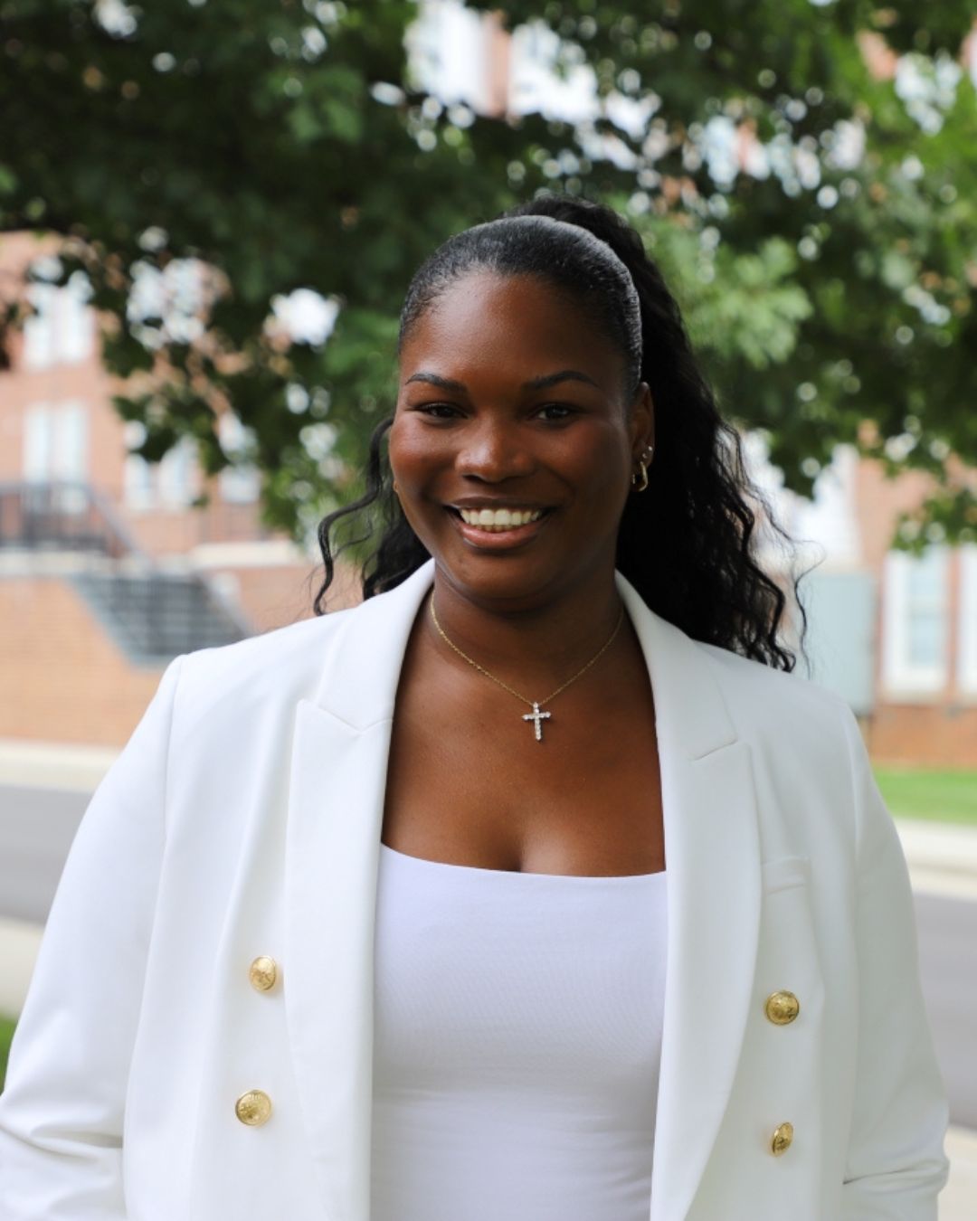 A woman wearing a white blazer and white shirt with a ponytail is smiling standing in front of a tree.