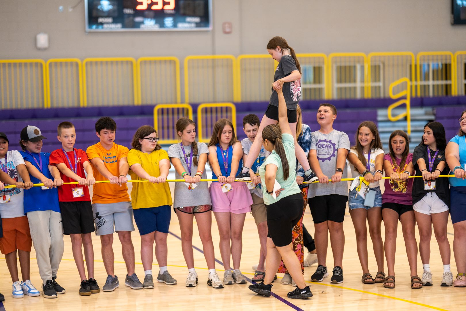 A group of around 20 middle school students stands side by side in a gymnasium, each holding onto a long yellow-and-green pole positioned at chest height. In the center of the group, a girl is carefully walking across the pole with the help of two adults—one woman holds her steady by the hand while another stands behind for support. The activity appears to be a trust-building or team exercise, likely part of a summer camp or school program. The students wear casual athletic clothes, name tags, and colorful lanyards. The gym features purple bleachers and yellow railings in the background, with a scoreboard visible overhead. The atmosphere is active, cooperative, and supportive.