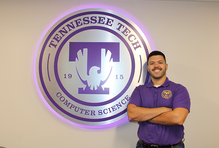 Computer science student stands next to computer science seal sign, smiling with his arms crossed