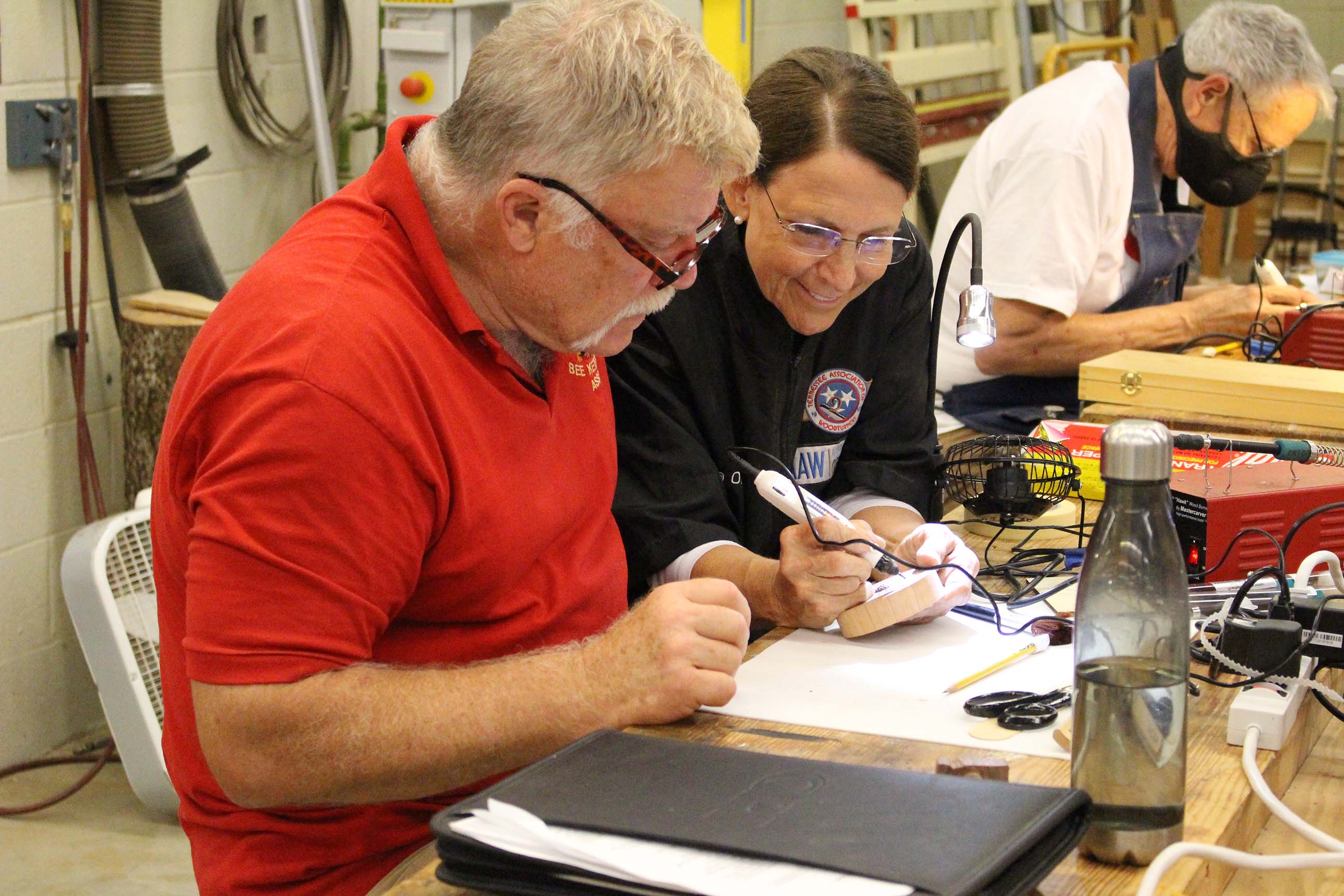 A woman showing a man in a red shirt how to do a craft
