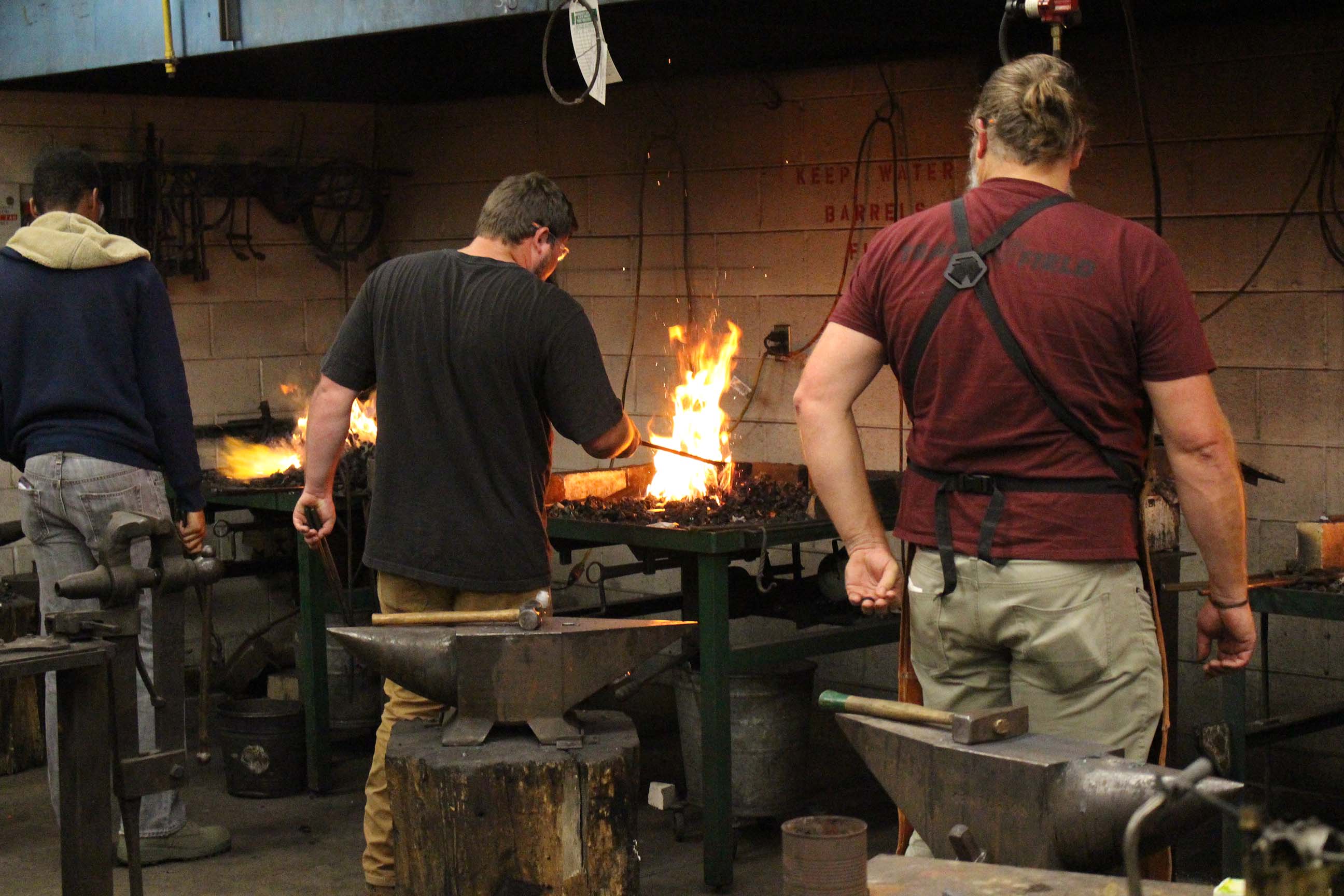 Three men heating metal in fires beside anvils