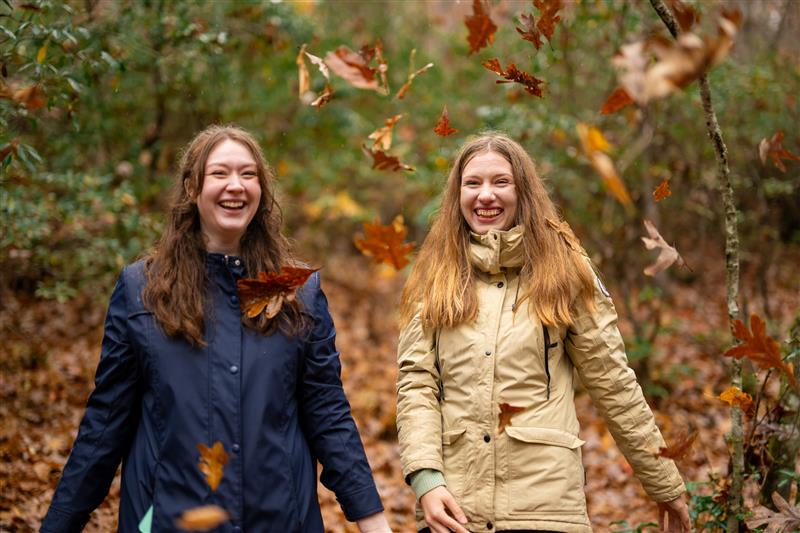 Two students hiling a trail while laughing and fall leaves are falling around them.