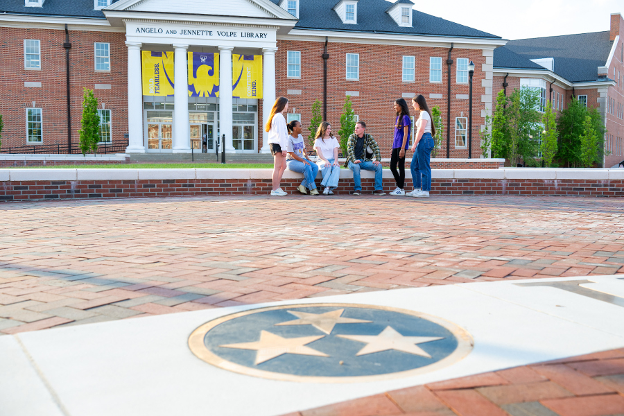 Photo of students hanging out in front of the library on the low brick ledge.