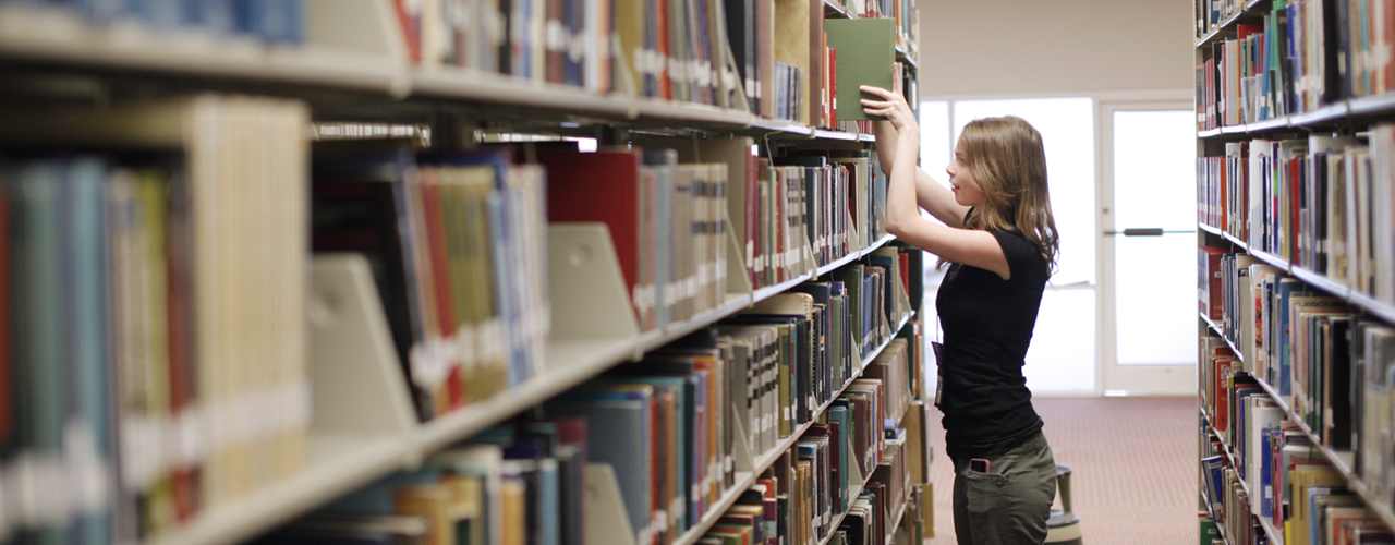 A female library student worker with brown shoulder-length hair is re-shelving a library book in the stacks.