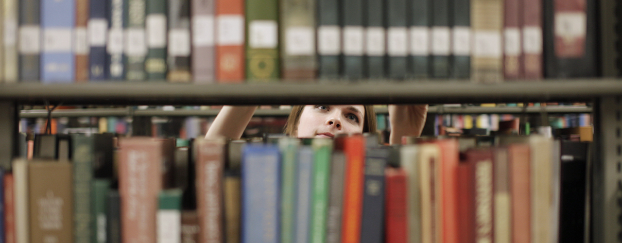 A library student worker's face is partially seen between two shelves on the opposite side of where the student is shelving a book.