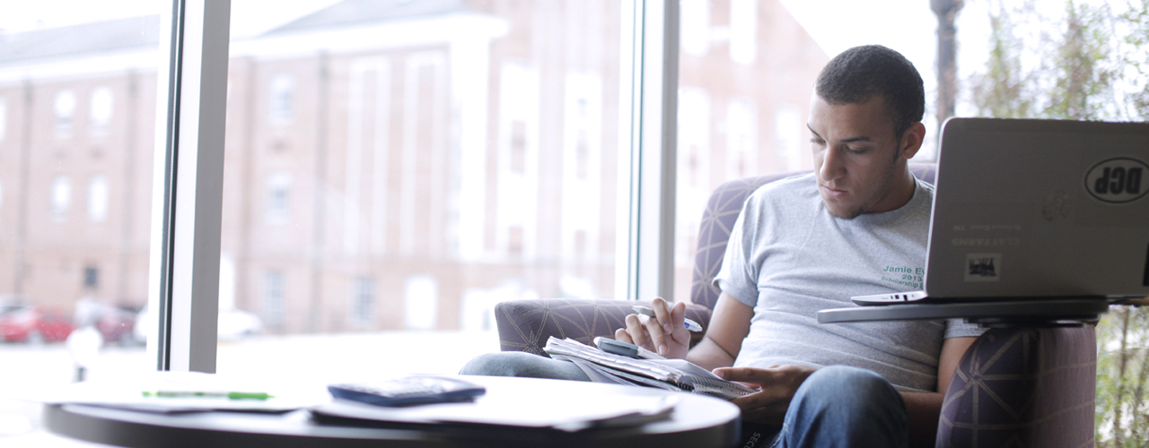 A male student in a gray t-shirt sits casually studying with a laptop on a study chair tray table.