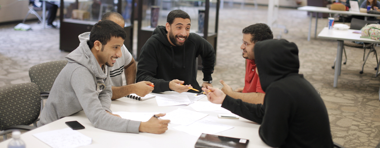 Four male students talk and study at a table in the library.