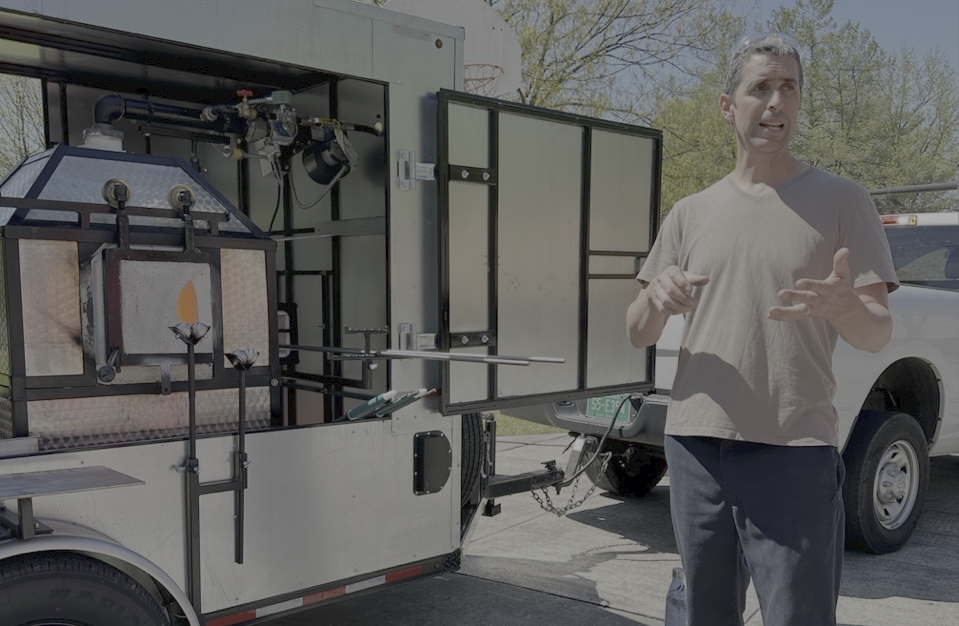 Damon MacNaught gestures in a t-shirt while standing in front of the Mobile GLass Unit. 