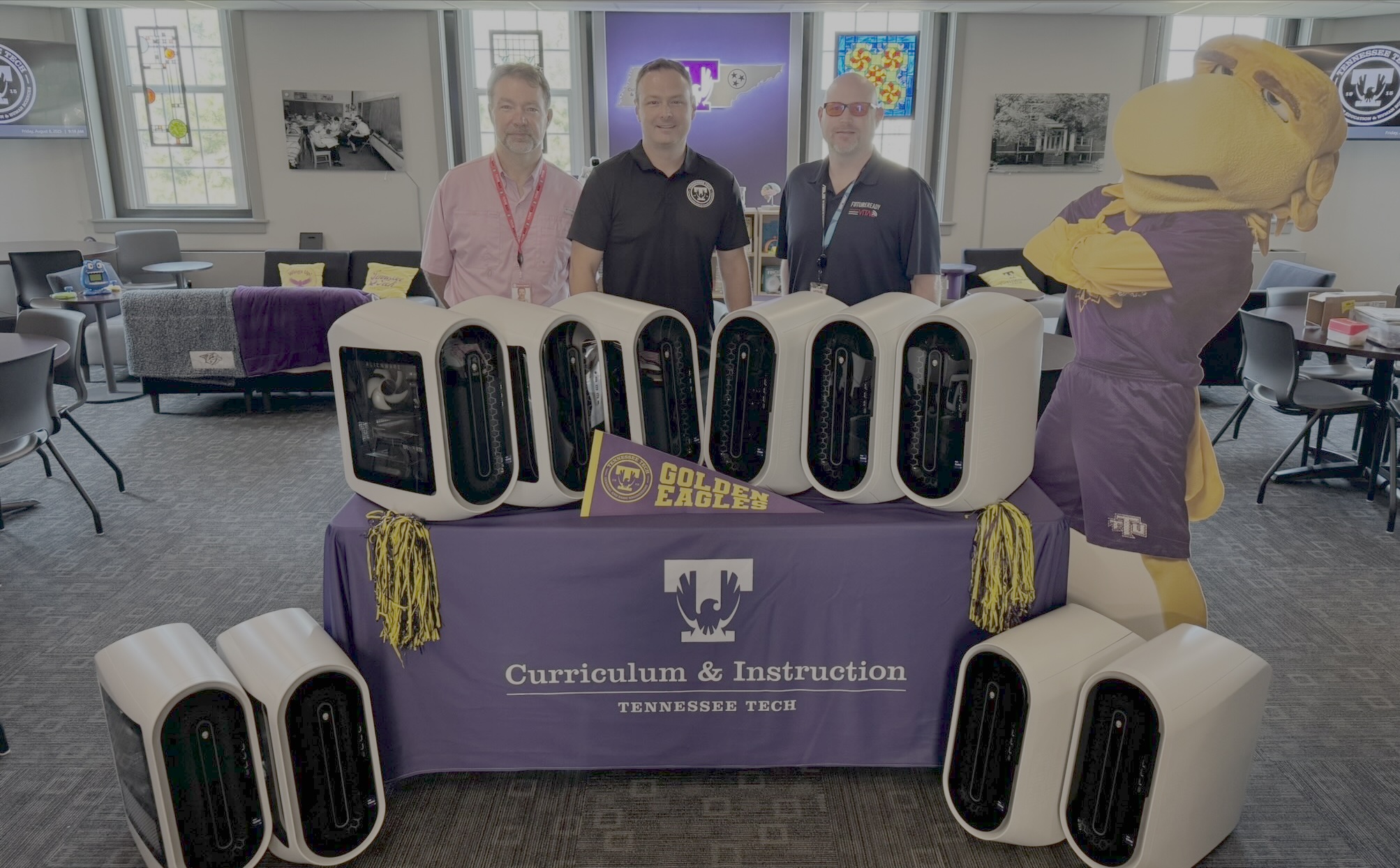 Three men stand at a table with 10 computers, a Tech pennant and a cutout of Awesome Eagle
