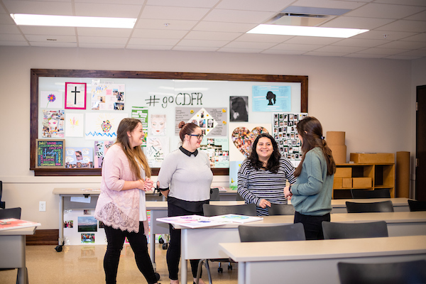 Photo of four students standing in front of a classroom talking.
