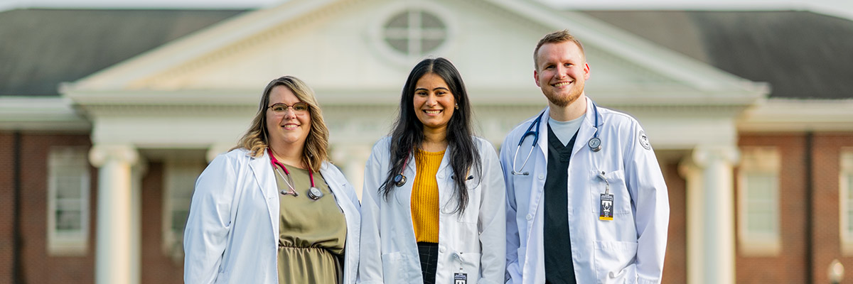 Three School of Nursing students standing in front of Bell Hall smiling at the camera as they pose with their scrubs on.