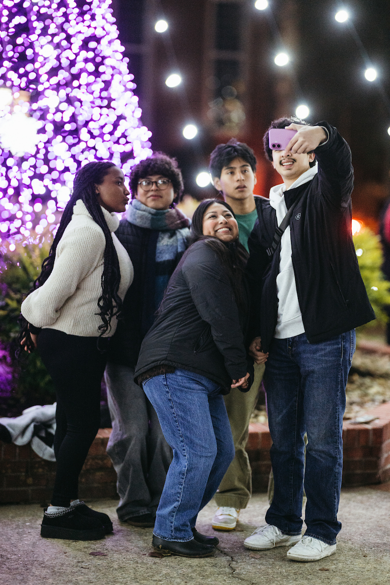 Five students taking a selfie outside with hoiday lights.