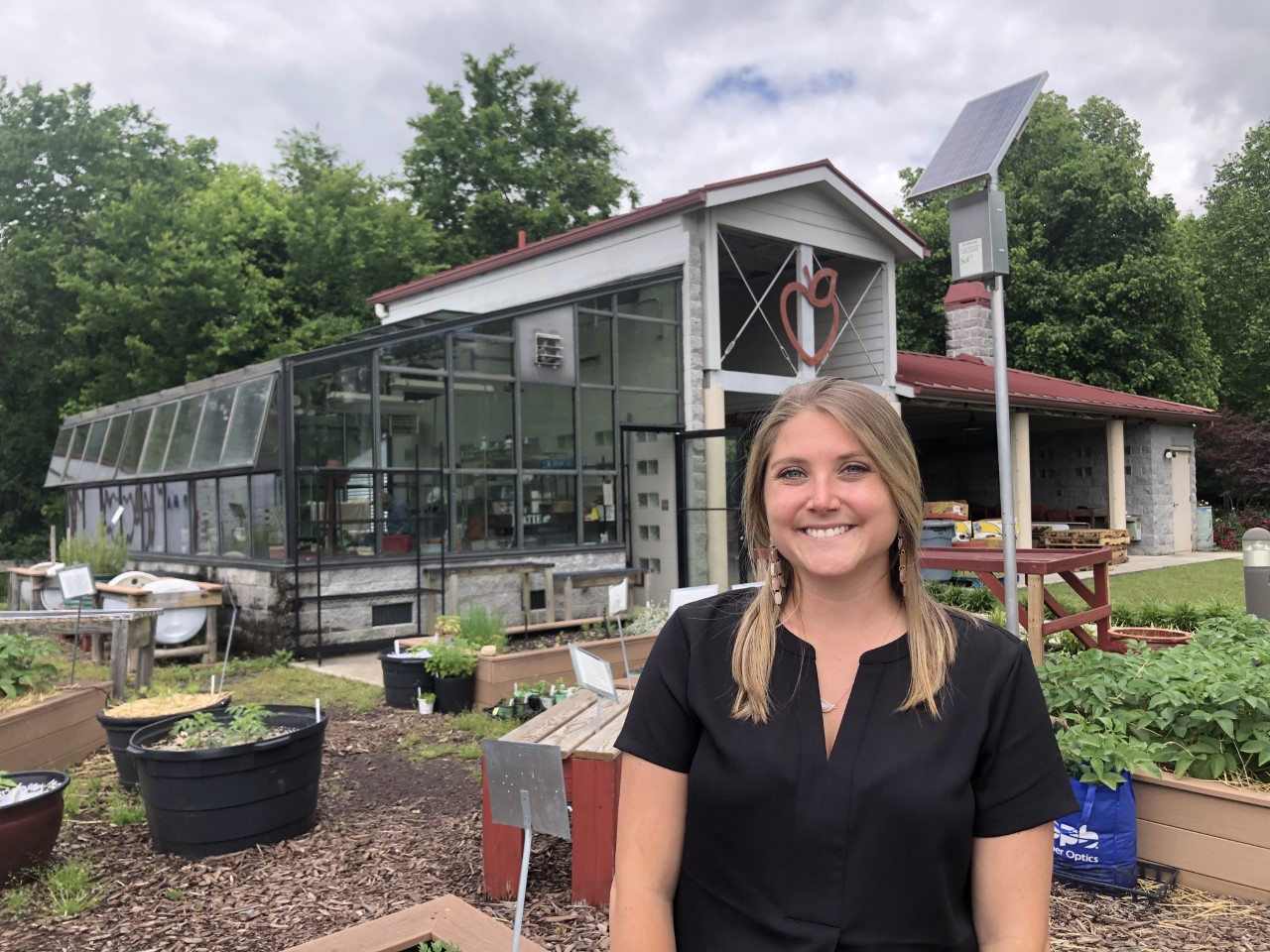 Photo of Abby Markus. Abby is standing in front of a garden and barn with a sign shaped like an apple. She has long blonde hair and a black shirt and is smiling.