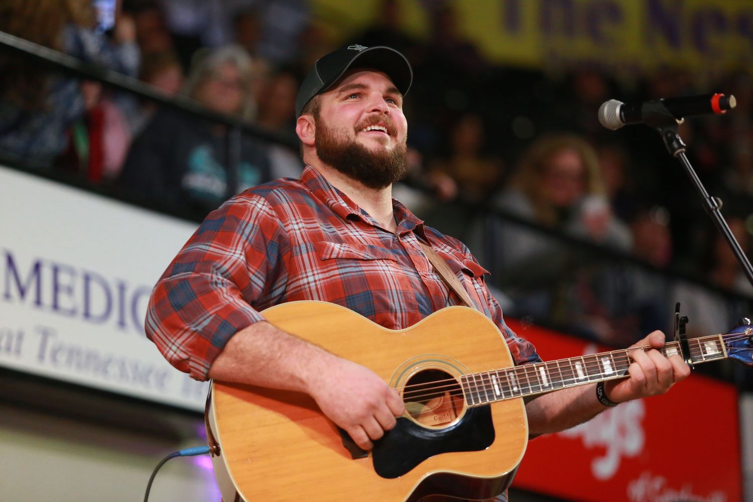 Jake Hoot is wearing a red flannel shirt and smiling as he play his guitar.