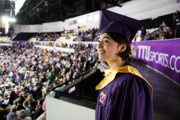 A female student stands in the upper part of Hooper Eblen Stadium in a purple graduation robe looking out at the stands packed with onlookers.