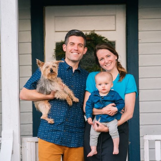 The Waugh Family is standing on their front porch. Brent is holding their pet dog and Megan is holding baby Willow.