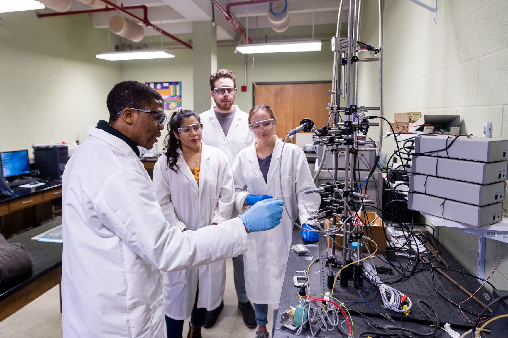 A group of students listen to a colleague discuss a lab apparatus.