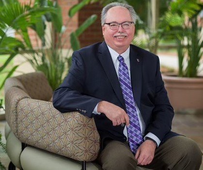 A photo of Mr. Ray smiling in a suit with a purple tie and sitting on a sofa in front of potted plants.