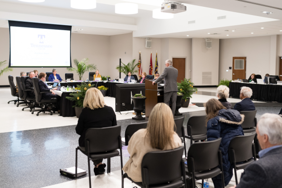 Board of Trustees sitting around a table with Dr. Oldham at a podium. There are people sitting in the foreground observing the meeting.