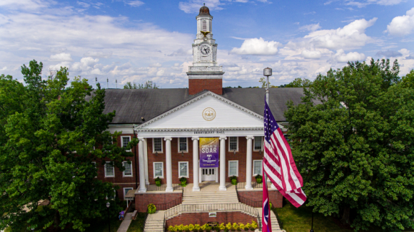 Aerial view of Derryberry Hall with a bright blue sky and white clouds. The American flag is in the foreground of the photo.