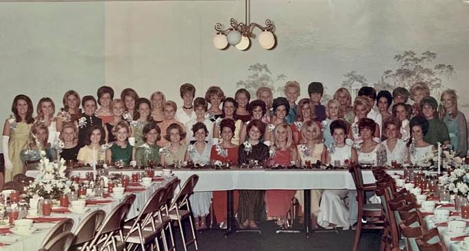 A group portrait from the 60s - young women with formal dresses and corsages smile behind banquet tables
