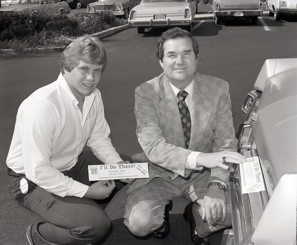 Two gentlemen placing a bumper sticker advertising homecoming on a car.