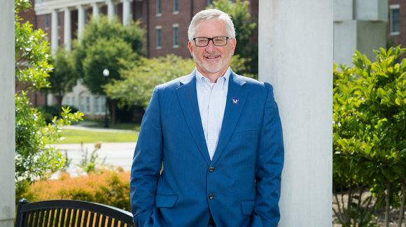 A photo of President Phil Oldham in a blue suit standing in front of the pergola on the Centennial Plaza