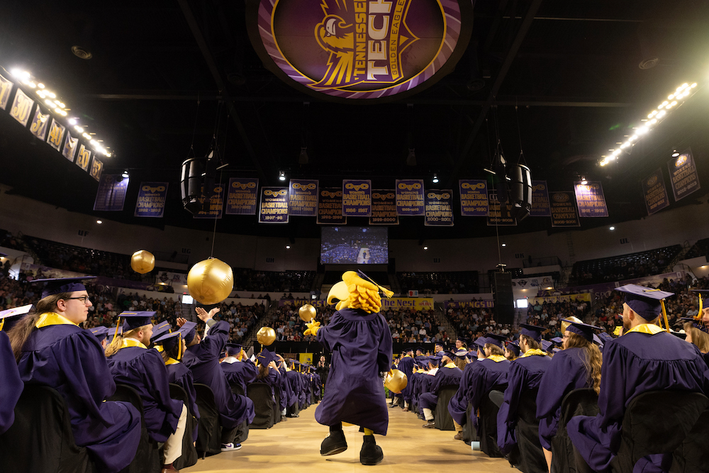 Awesome Eagle walks down the center aisle at commencement wearing graduation regalia. On either side of the aisles are students seated in regalia.