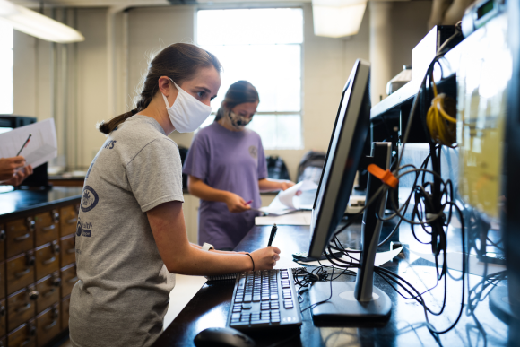 Two female students wearing face masks sit across from each other in a lab.